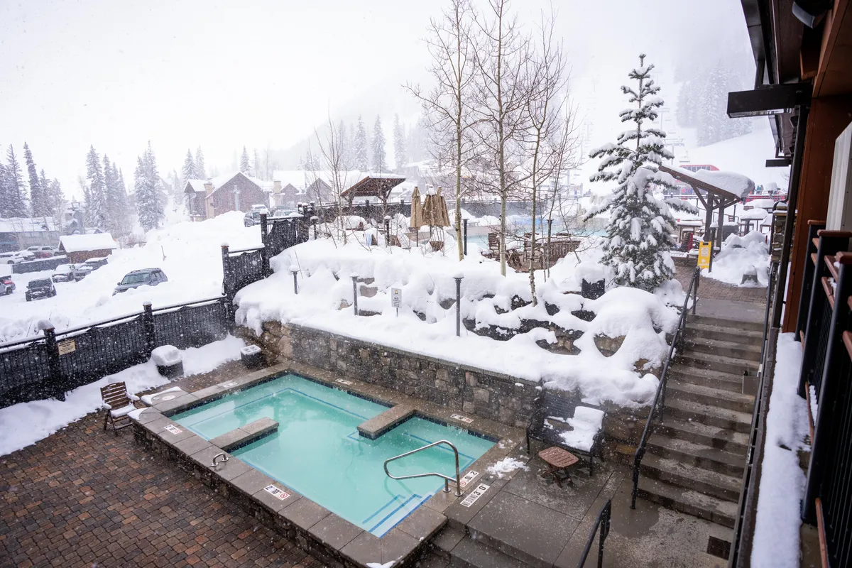 A serene outdoor hot tub set amid snow-covered trees and a rustic stone wall, with steps leading down. The ambiance is tranquil and inviting, perfect for relaxation during winter, with the allure of stargazing while enveloped in warmth.