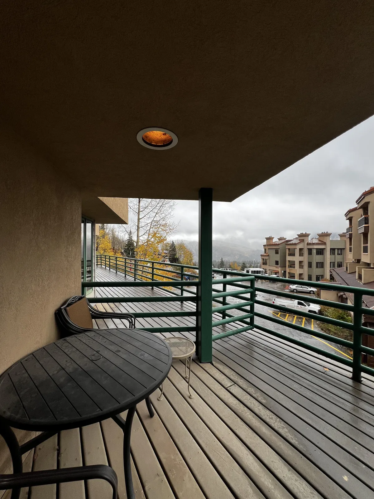 A relaxed exterior scene showing a private deck with a table and chairs, overlooking trees and surrounding buildings. The deck invites guests to step outside for a meal, with mountain views implied beyond the railing.