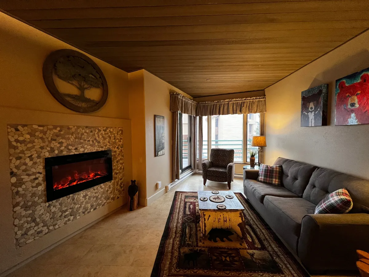 A warm, welcoming interior scene featuring a plush gray sofa set around a modern fireplace. Large windows with curtains frame the space, inviting natural light and hinting at potential mountain views. The artwork featuring bears and a wooden coffee table complete the cozy, open living area perfect for relaxing after a day outdoors.