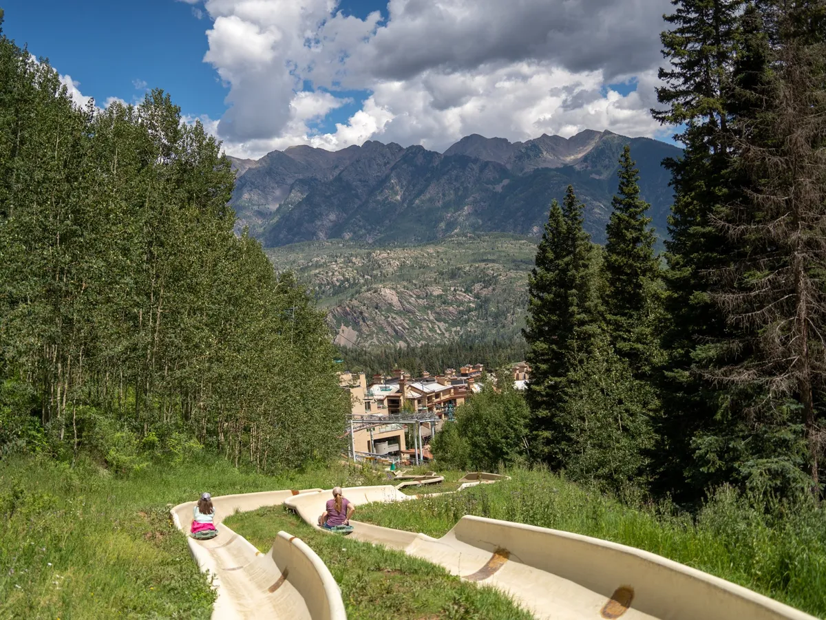 An exterior scene showing a vibrant mountain backdrop with people enjoying the outdoor sliding track. The lush landscape sets a joyful, adventurous mood, highlighting both the proximity to nature and the array of outdoor activities available nearby.