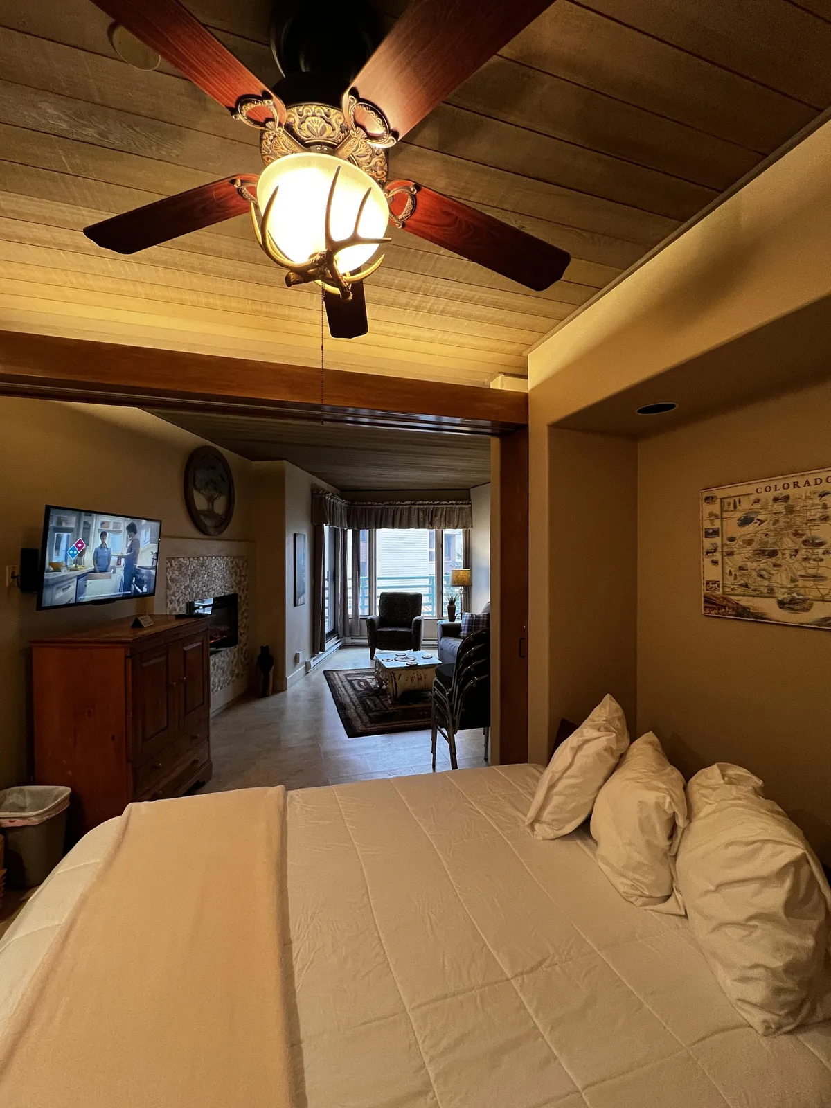 A bright, comfortable bedroom scene with a bed dressed in fresh white linens, a ceiling fan with light, wooden dresser, TV, fireplace, and a seating area with two chairs. The arrangement showcases both sleeping and relaxing areas, reinforcing the studio's flexibility and comfort.