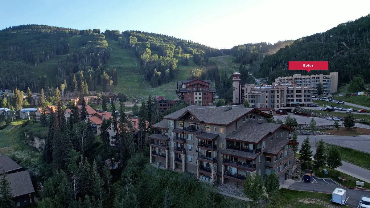 A wide shot of the resort's residential buildings nestled among greenery, with a visible parking area in the foreground and mountains in the background. The image conveys convenience of access and a sense of arrival in a scenic alpine environment.