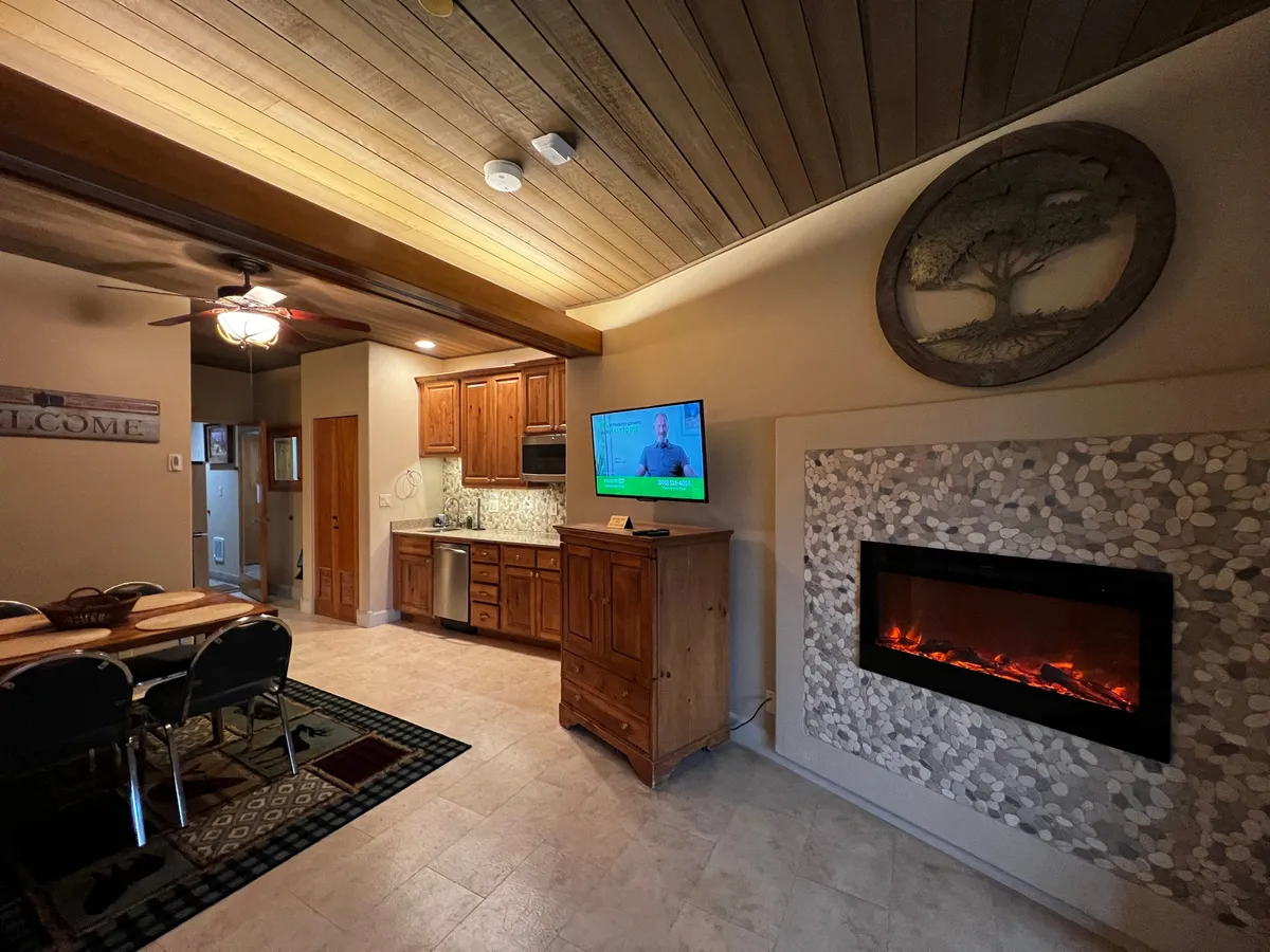 A cozy interior scene featuring a dining table set near the efficiency kitchen, with wooden cabinetry, a warm fireplace, and a television. The space is ideal for enjoying meals and intimate gatherings, while the kitchen equipment and cabinetry offer practical convenience for food preparation.