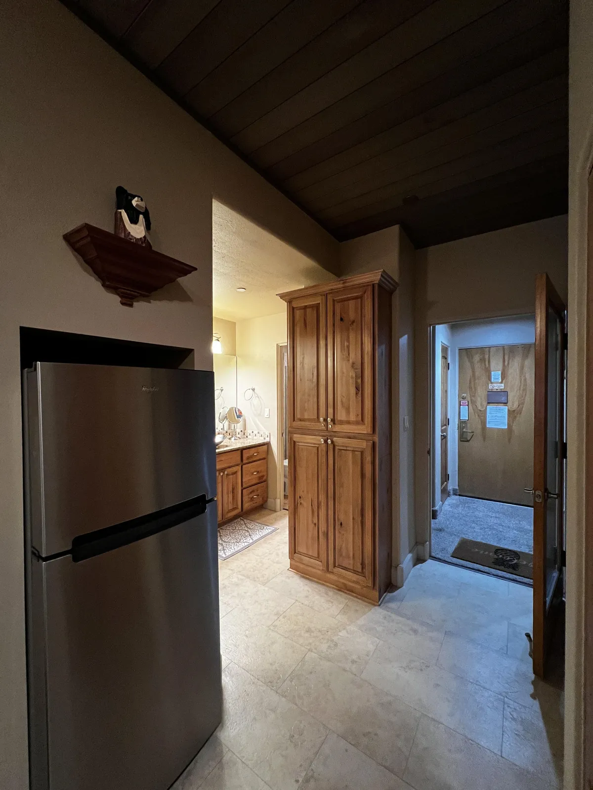 A view of the kitchen area featuring a refrigerator, wooden cabinetry, and a decorative item on the shelf. The lighting creates an inviting atmosphere ideal for preparing a morning coffee or an intimate meal.