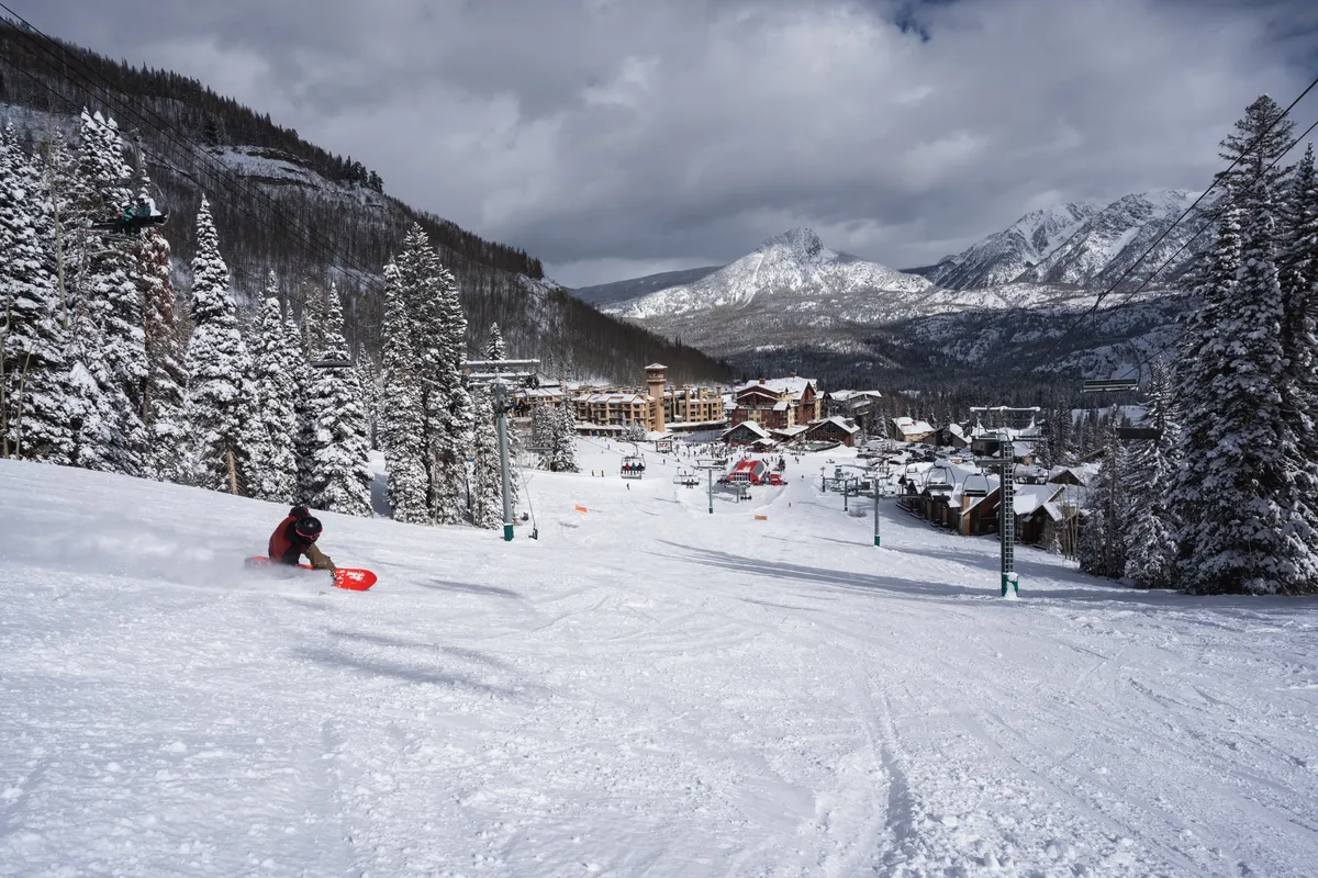 This image captures a skier descending a snowy slope with ski lifts rising in the background, surrounded by snow-covered trees and a mountain range, with resort buildings visible at the base. The scene is dynamic and illustrates both the thrill of winter sports and the proximity of accommodations to the action.