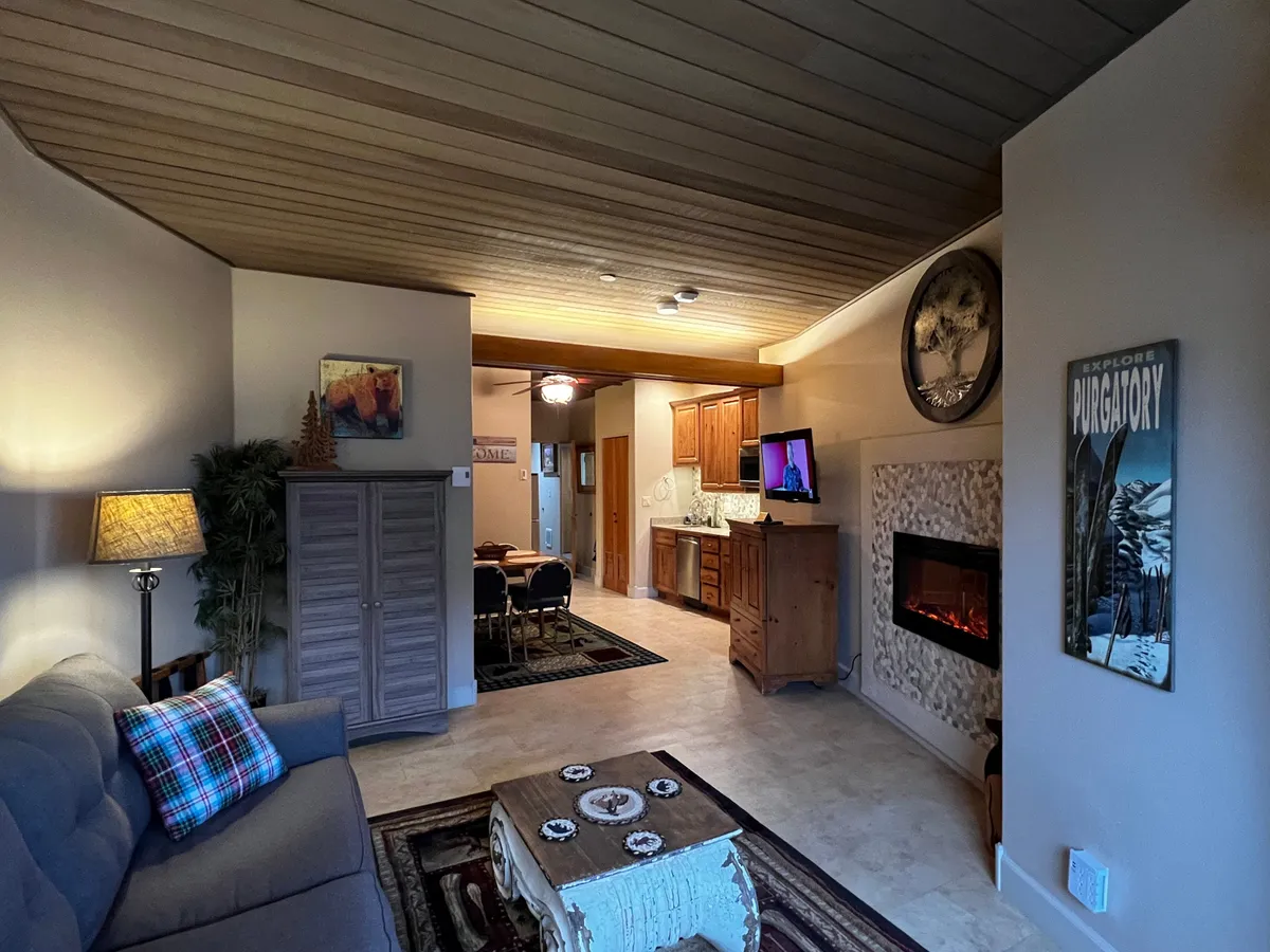 The image showcases a cozy studio living area featuring a comfortable sofa draped with a plaid throw, a crackling fireplace, a wooden dining table perfect for two, and stylish decorative wall art. The scene feels warm and inviting, ideal for relaxing after a day on the slopes or enjoying an intimate meal by the fire.