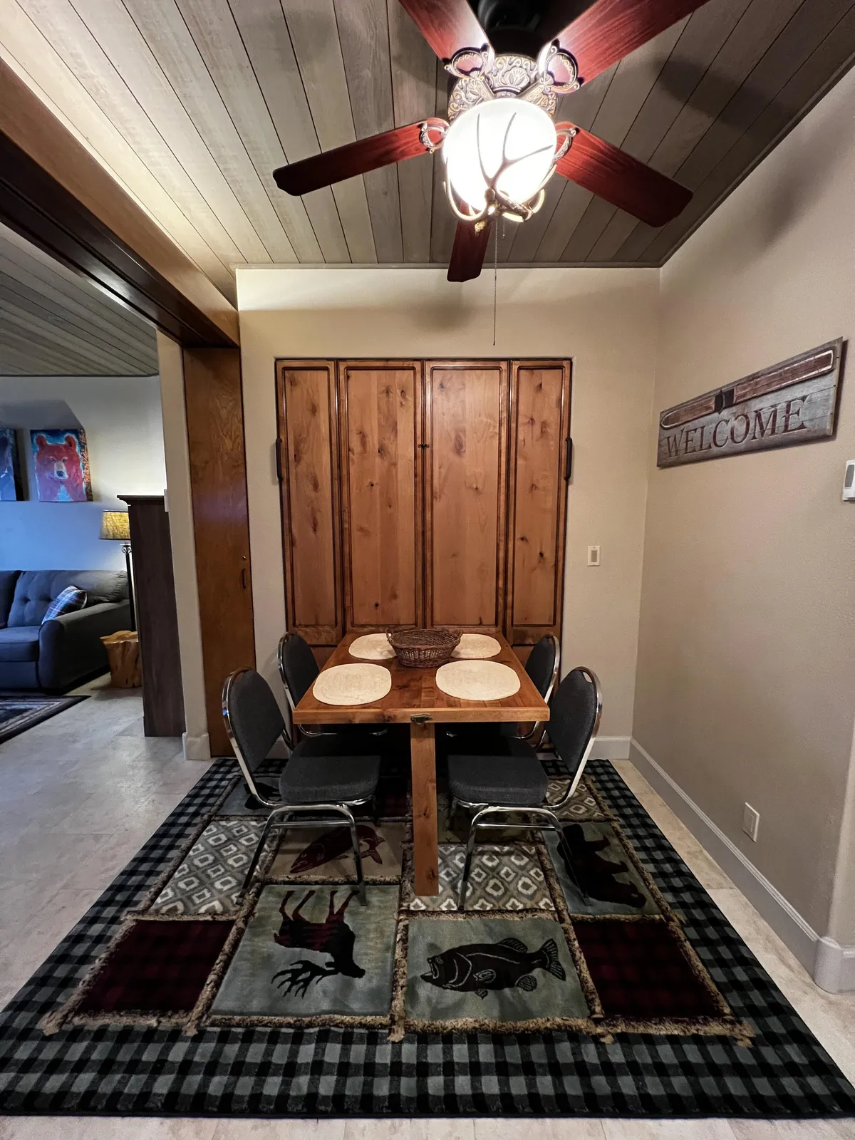 This interior scene features a welcoming dining area with a wooden table set, warm cabinetry, and a visible 'welcome' sign. The ceiling fan above hints at comfort for all seasons, while the overall atmosphere is one of casual, communal gathering.